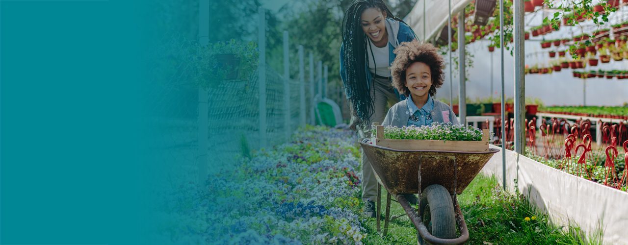 A happy child being pushed in a wheelbarrow filled with flowers by a smiling woman.